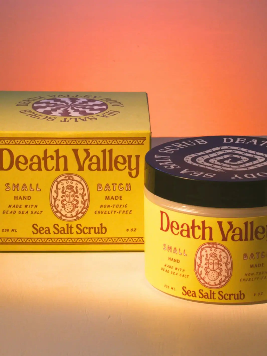 The yellow jar and outer box for Death Valley Nails Dead Sea Salt Scrub - Lemon + Sage + Peppermint + Earth sit side-by-side under warm lighting with a pink gradient background | Twentyseven Toronto