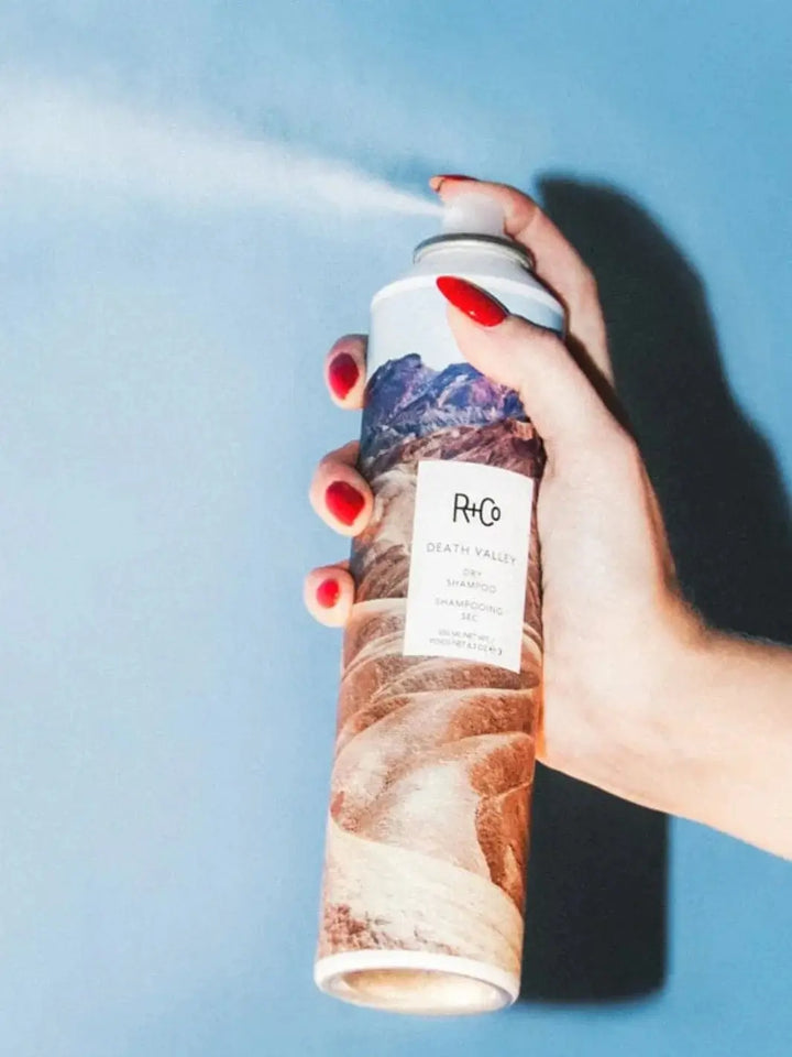A hand with red fingernails sprays a fine mist from a can of R+Co SPIRITUALIZED Wet Dry Shampoo against a solid blue background | Twentyseven Toronto