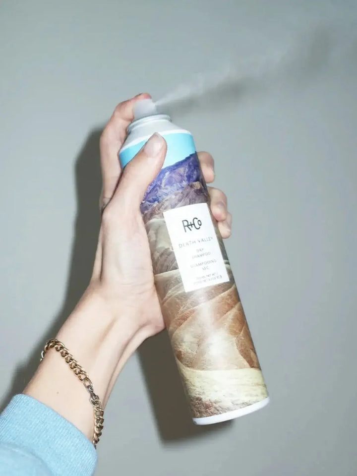 A hand wearing a gold chain bracelet presses the nozzle of a can of R+Co SPIRITUALIZED Wet Dry Shampoo to release a continuous mist | Twentyseven Toronto