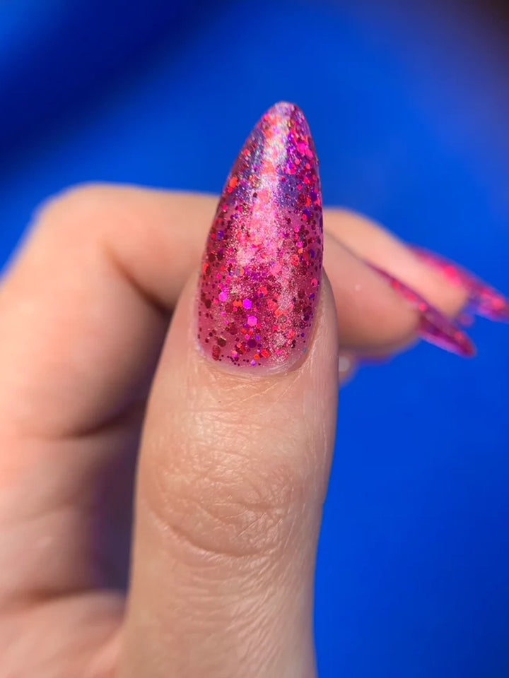 Close-up of pink multichrome glitter polish on a long almond-shaped nail against a blue background | Twentyseven Toronto