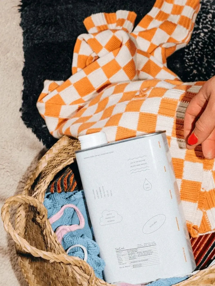 A white tin of Dedcool Dedtergent 01 "Taunt" sits inside a woven laundry basket next to an orange and white checkered sweater | Twentyseven Toronto