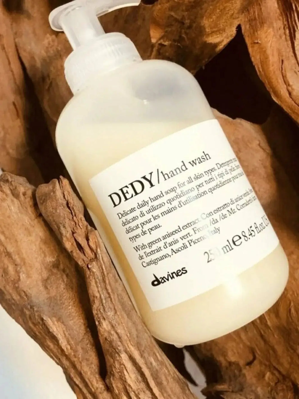 A translucent plastic pump bottle of Davines DEDY Hand Wash is nestled at an angle against pieces of textured brown driftwood | Twentyseven Toronto