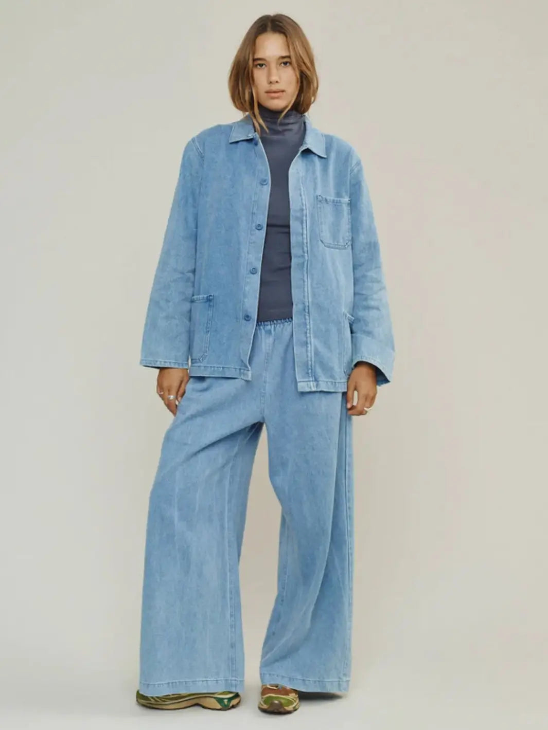 A full-length studio portrait features a model wearing the unbuttoned Jungmaven Denim Olympic Jacket in Light styled as a matching set with wide-leg denim trousers and a dark grey turtleneck | Twentyseven Toronto