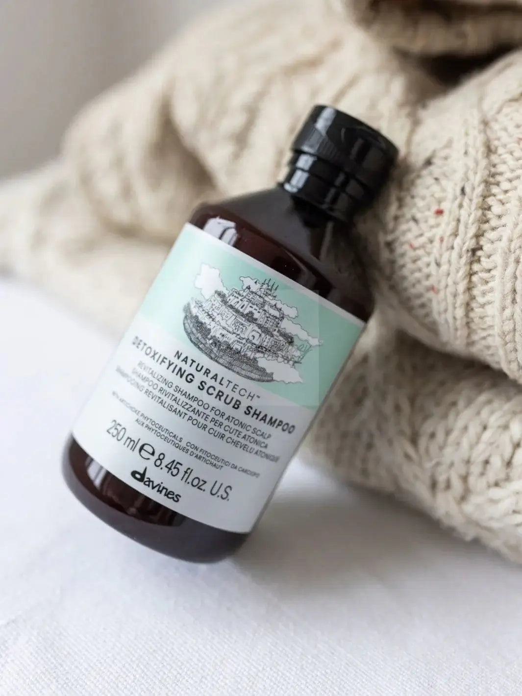A bottle of Davines DETOXIFYING Scrub Shampoo rests against a chunky, cream-colored knit sweater | Twentyseven Toronto