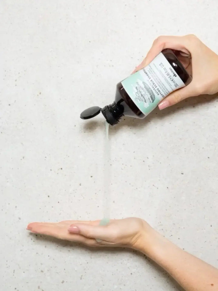 A light green liquid pours from a bottle of Davines DETOXIFYING Scrub Shampoo into an open hand against a speckled white wall | Twentyseven Toronto