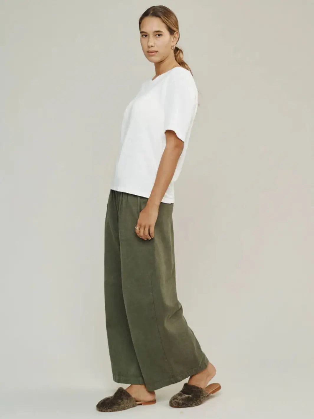 A side profile shot captures a model wearing a classic white t-shirt styled with the wide-leg Jungmaven Disco Twill Pant - Olive Green and fuzzy brown slippers | Twentyseven Toronto
