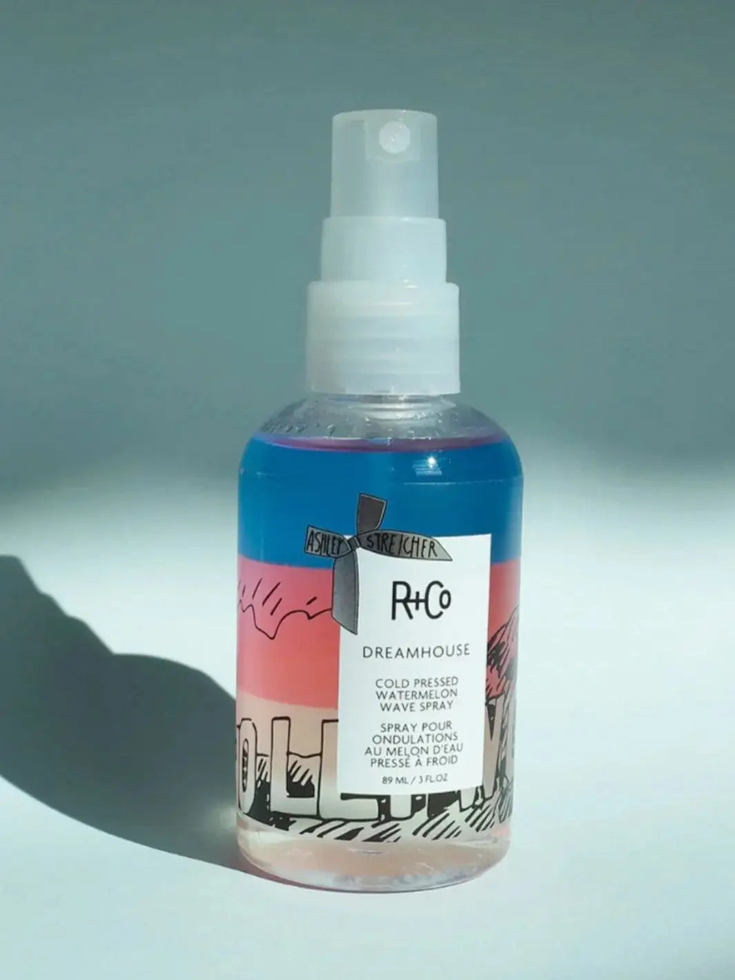 A bottle of R+Co DREAMHOUSE Cold Pressed Watermelon Spray featuring a bi-phase blue and pink formula stands upright against a light blue background | Twentyseven Toronto
