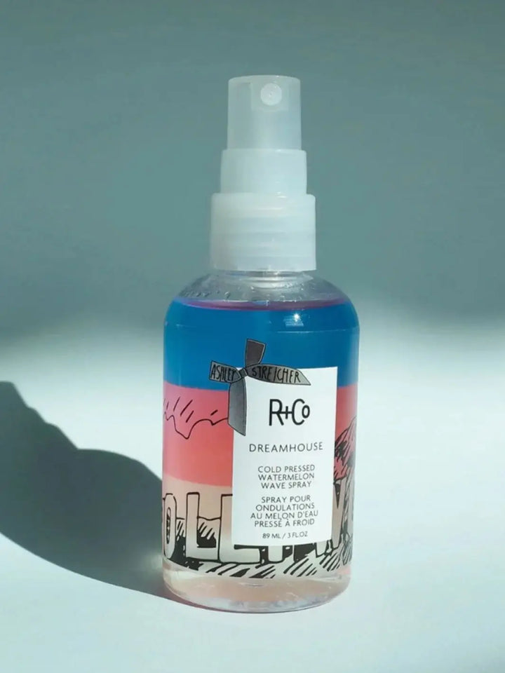 A bottle of R+Co DREAMHOUSE Cold Pressed Watermelon Spray featuring a bi-phase blue and pink formula stands upright against a light blue background | Twentyseven Toronto
