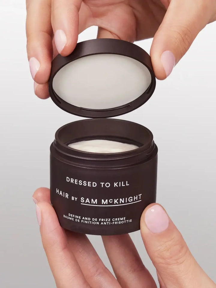 Two hands with pale pink nail polish opening the lid of a dark jar of Hair by Sam McKnight Dressed to Kill Define and Defrizz Creme to reveal the white cream inside | Twentyseven Toronto