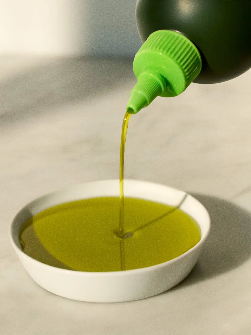 Green bottle of Graza 'Drizzle' pouring olive oil into a white bowl on a light surface | Twentyseven Toronto