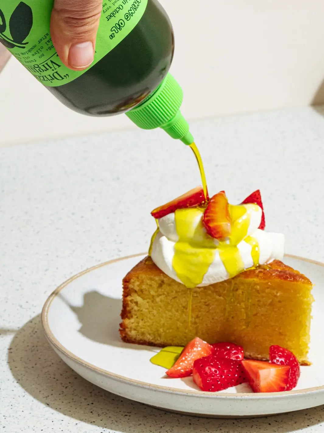 Sliced cake with strawberries and whipped cream being drizzled with a green bottle of Graza 'Drizzle' olive oil on a white plate | Twentyseven Toronto