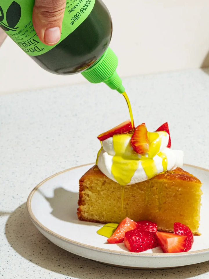 Sliced cake with strawberries and whipped cream being drizzled with a green bottle of Graza 'Drizzle' olive oil on a white plate | Twentyseven Toronto