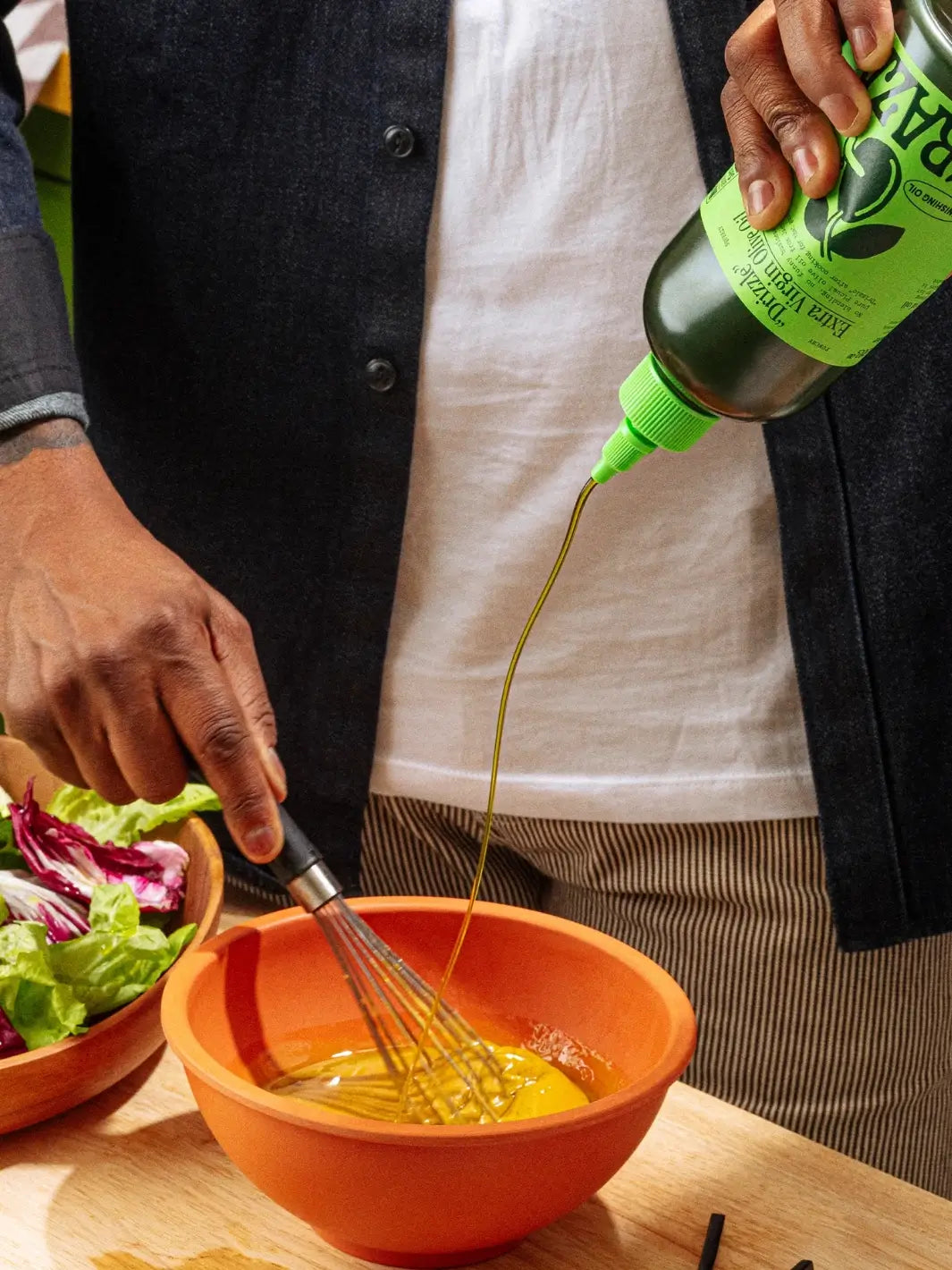 Person whisking eggs in an orange bowl with a green bottle of Graza 'Drizzle' olive oil being poured over them | Twentyseven Toronto