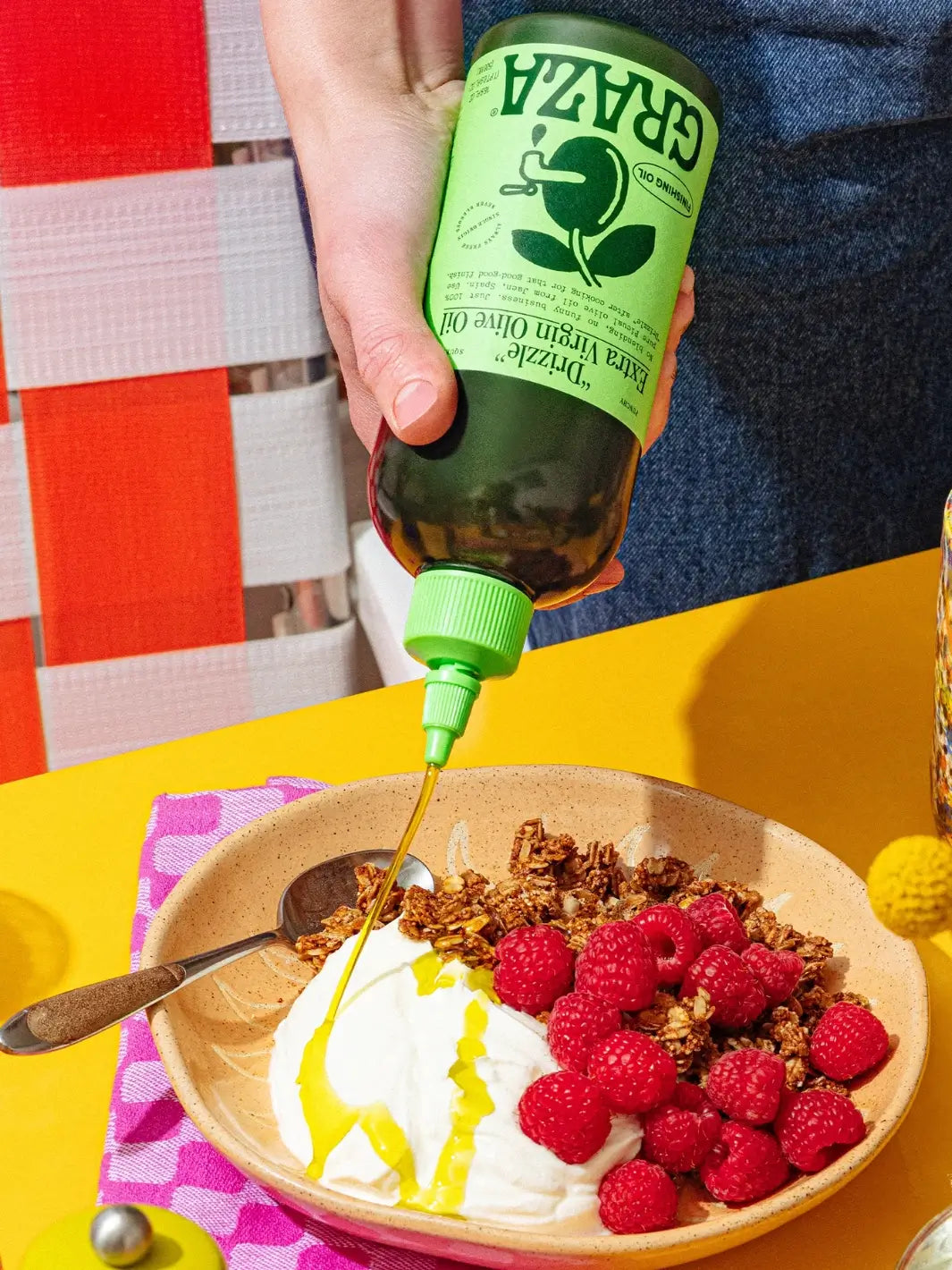 Person pouring Graza 'Drizzle' olive oil over a bowl of yogurt and raspberries on a colourful table | Twentyseven Toronto