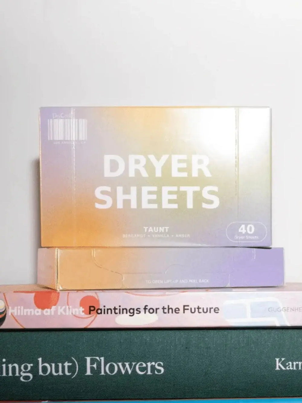 A box of Dedcool Dryer Sheets Taunt featuring an orange and purple gradient design is stacked neatly on top of two art books | Twentyseven Toronto