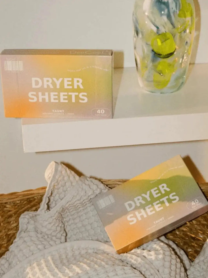 One box of Dedcool Dryer Sheets Taunt sits on a sleek white shelf next to a green glass vase, while another box rests below in a woven basket filled with a grey waffle-weave towel | Twentyseven Toronto