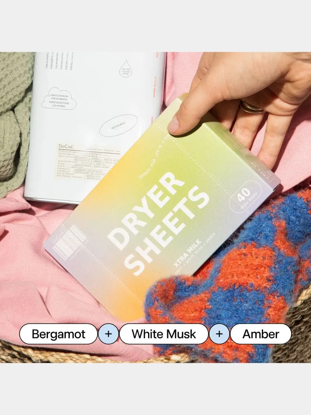A hand holds a box of Dedcool Dryer Sheets Xtra Milk inside a laundry basket filled with colorful clothing and a matching detergent tin, with ingredient labels displayed at the bottom | Twentyseven Toronto