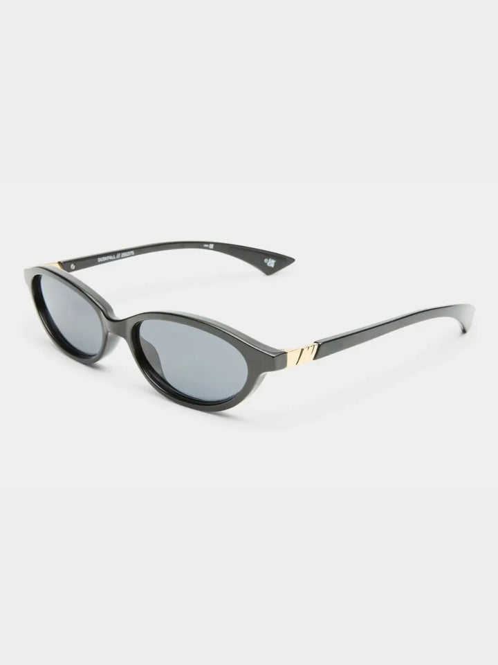 A three-quarter studio product shot of the Le Specs Duskfall - Black Smoke Mono sunglasses showcasing their glossy black frame, dark lenses, and textured gold temple accents | Twentyseven Toronto