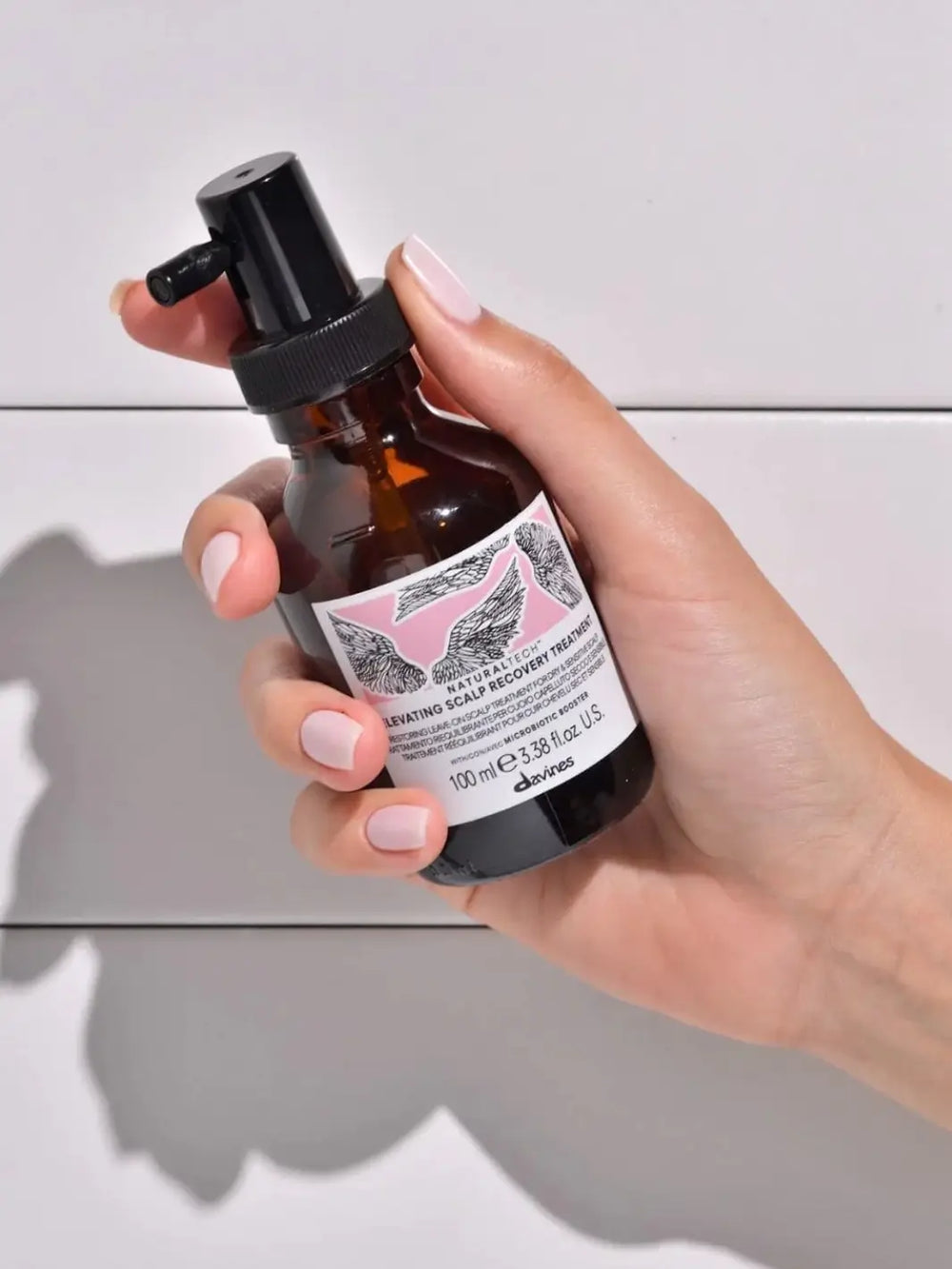 A hand with light pink nail polish holds a pump bottle of Davines ELEVATING Scalp Recovery Treatment against a white background | Twentyseven Toronto