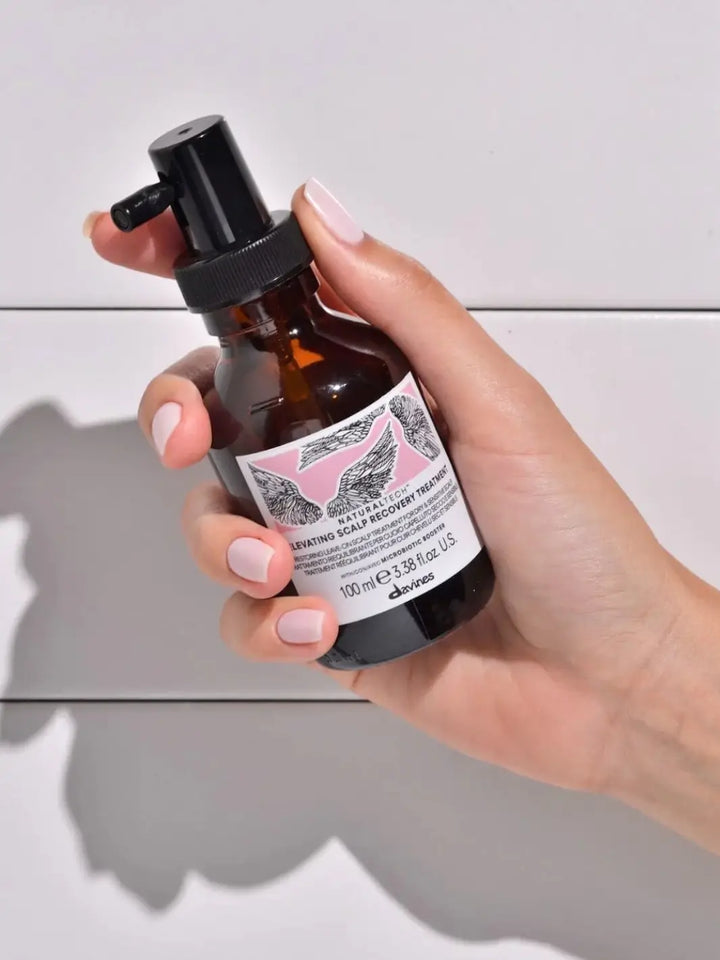 A hand with light pink nail polish holds a pump bottle of Davines ELEVATING Scalp Recovery Treatment against a white background | Twentyseven Toronto