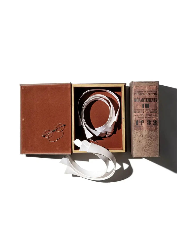 Top-down view of an open antique-style book box acting as a Decoration Tray / Oval from Puebco, containing a coiled white ribbon and wire-rimmed spectacles on a velvet lining | Twentyseven Toronto