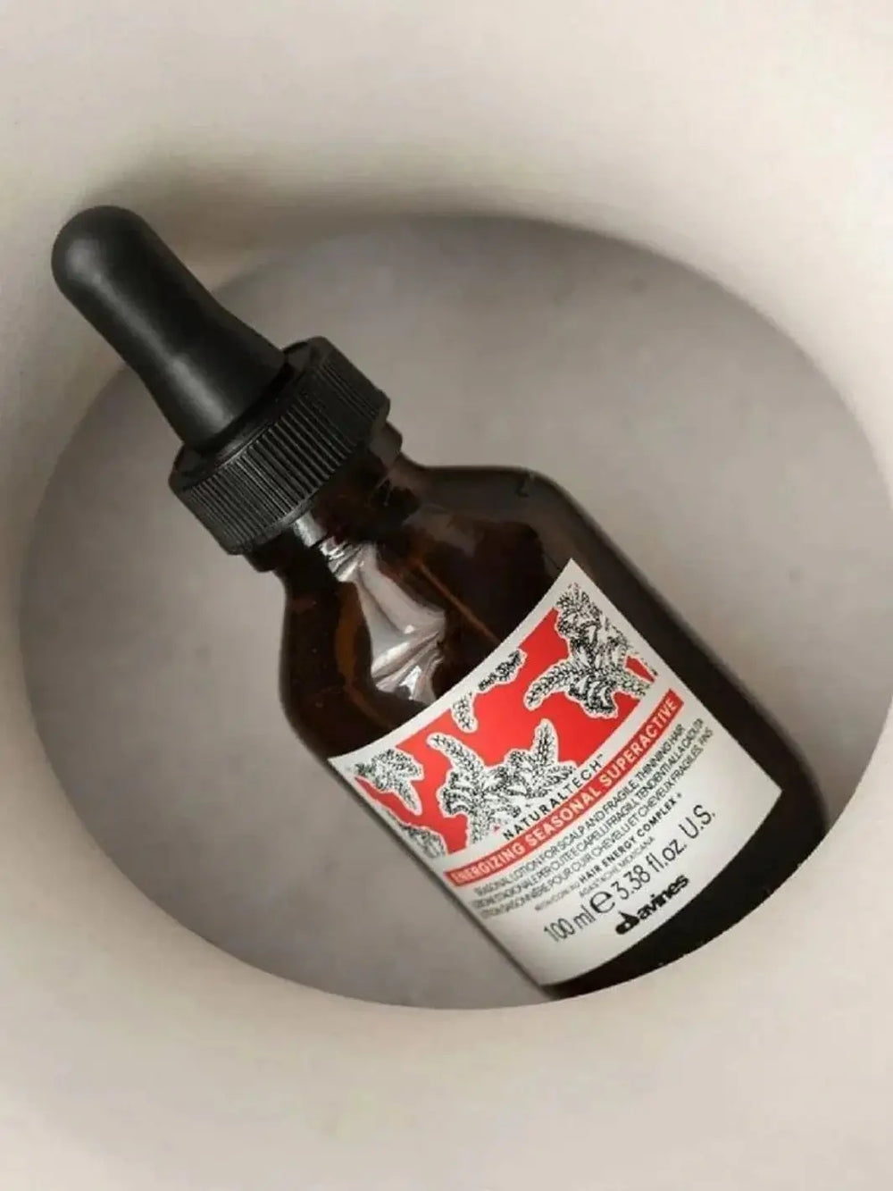 An amber glass dropper bottle of Davines ENERGIZING Seasonal Superactive is tilted inside a smooth, circular grey vase | Twentyseven Toronto