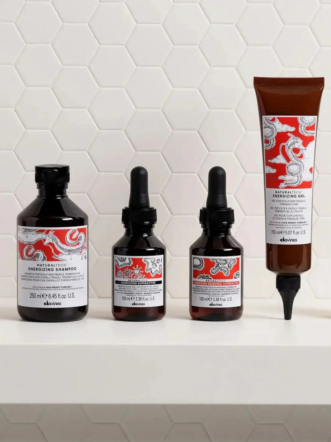 A bottle of Davines ENERGIZING Superactive Anytime Serum stands in a row with a matching shampoo, seasonal treatment, and hair gel against a white tile background | Twentyseven Toronto