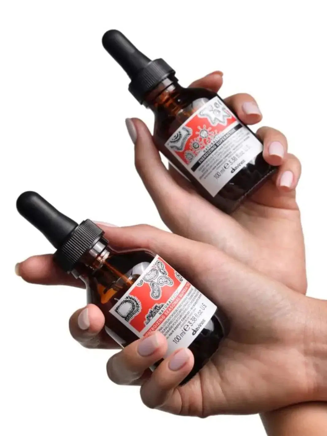 A person holds a bottle of Davines ENERGIZING Superactive Anytime Serum in one hand and a seasonal treatment bottle in the other against a plain white background | Twentyseven Toronto