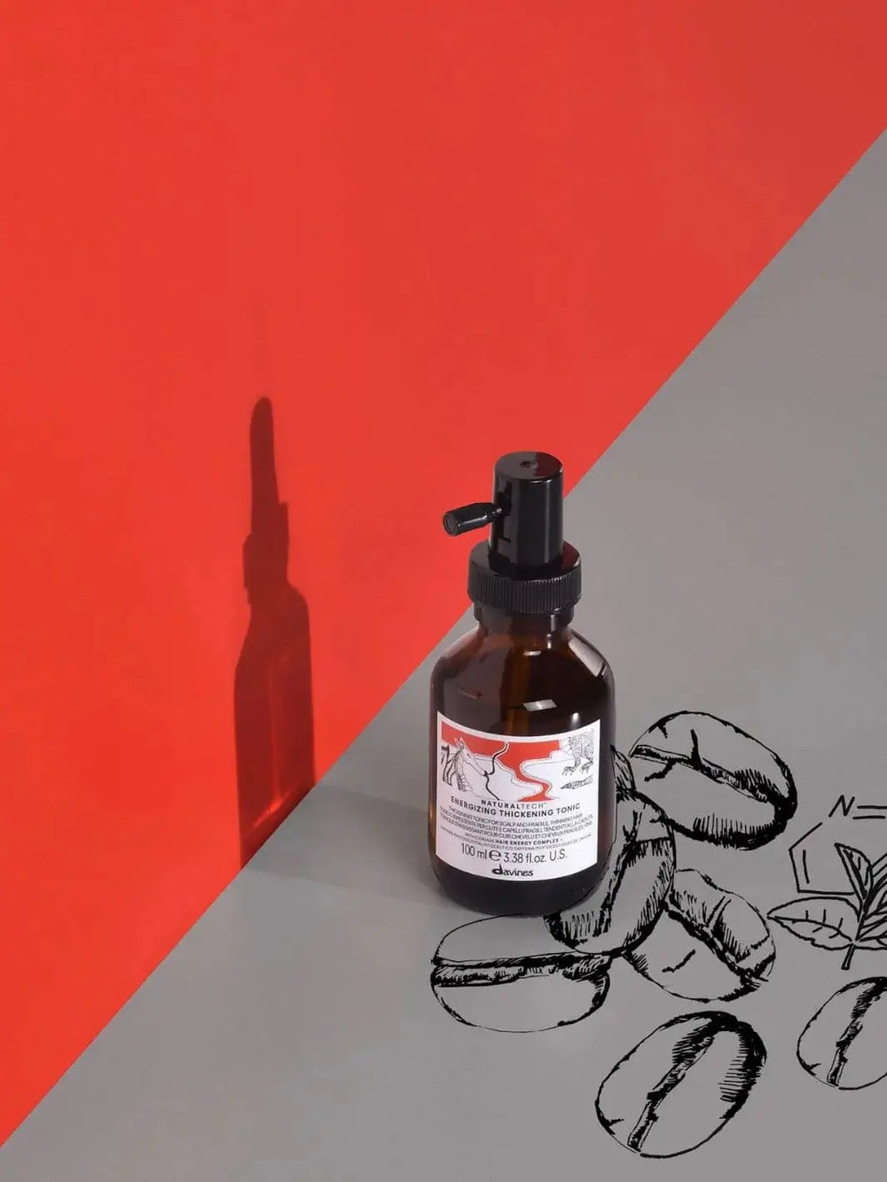 A bottle of Davines ENERGIZING Thickening Tonic sits on a grey surface decorated with drawings of coffee beans, casting a shadow against a bright red backdrop | Twentyseven Toronto