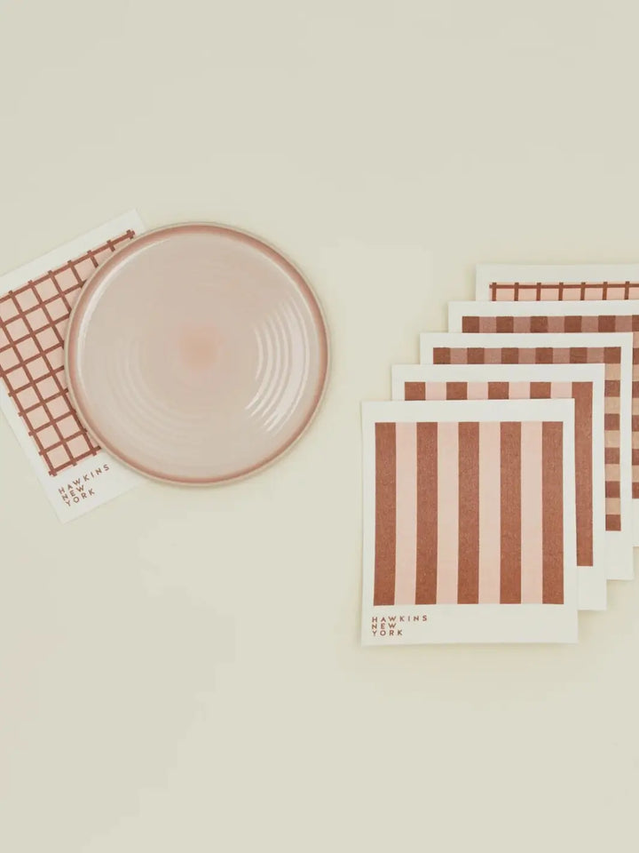 A flat lay features a pink ceramic plate resting on a windowpane sponge cloth alongside a stacked bundle of coordinating patterns from the Essential Swedish Dishcloth in Blush/Terracotta (Set of 6) from Hawkins New York | Twentyseven Toronto