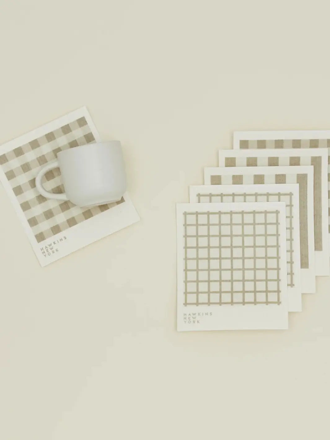 A flat lay features a white ceramic mug resting on a plaid sponge cloth alongside a stacked bundle of coordinating patterns from the Essential Swedish Dishcloth in Ivory/Flax (Set of 6) from Hawkins New York | Twentyseven Toronto