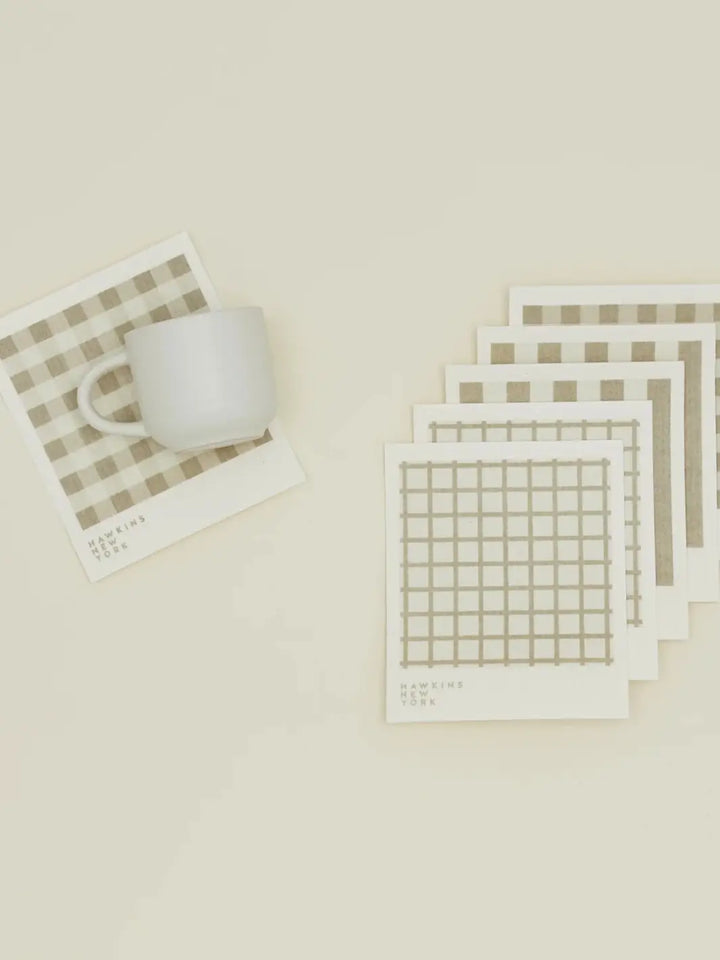 A flat lay features a white ceramic mug resting on a plaid sponge cloth alongside a stacked bundle of coordinating patterns from the Essential Swedish Dishcloth in Ivory/Flax (Set of 6) from Hawkins New York | Twentyseven Toronto
