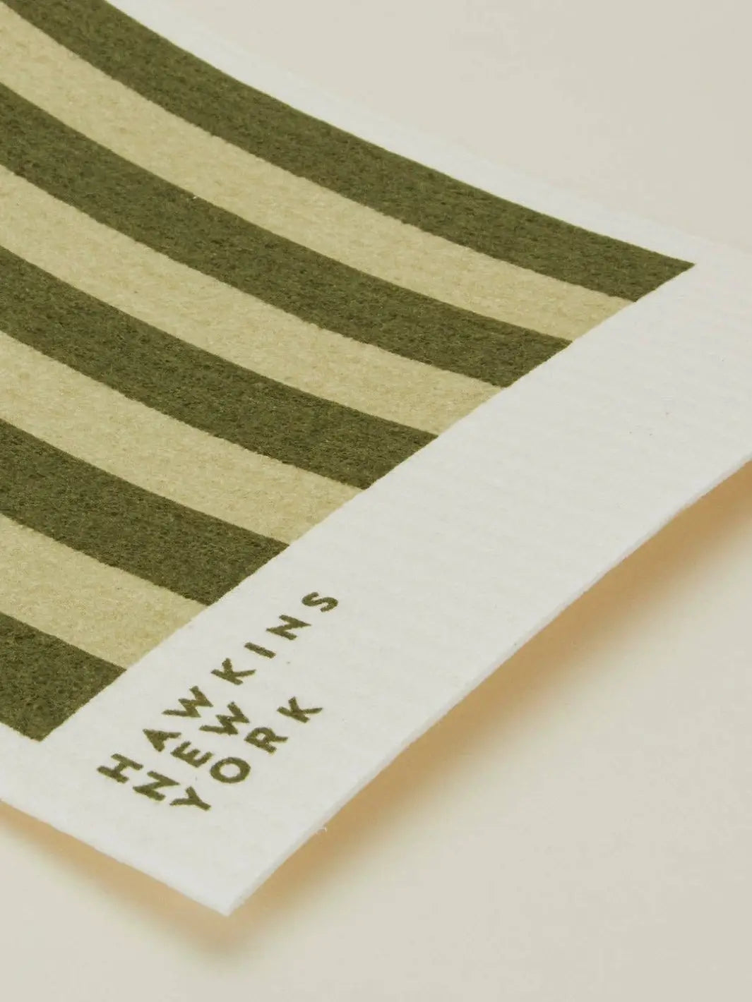 A close-up shot focuses on the texture and brand logo of an olive green and cream striped sponge cloth from the Essential Swedish Dishcloth in Sage/Olive (Set of 6) from Hawkins New York | Twentyseven Toronto