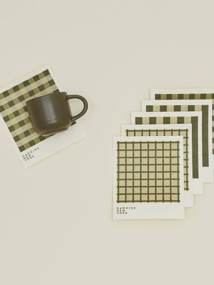 A flat lay features a dark ceramic mug resting on a plaid sponge cloth alongside a stacked bundle of coordinating patterns from the Essential Swedish Dishcloth in Sage/Olive (Set of 6) from Hawkins New York | Twentyseven Toronto