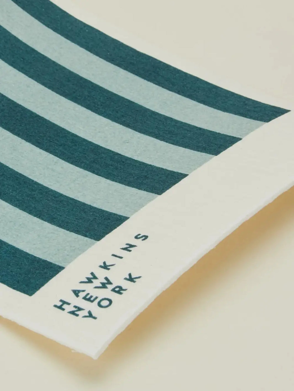 A close-up shot focuses on the texture and brand logo of a sky blue and dark blue striped sponge cloth from the Essential Swedish Dishcloth in Sky/Peacock (Set of 6) from Hawkins New York | Twentyseven Toronto