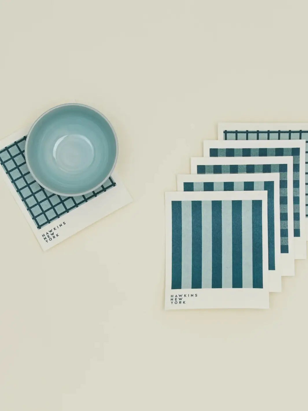 A flat lay features a light blue ceramic bowl resting on a windowpane sponge cloth alongside a stacked bundle of coordinating patterns from the Essential Swedish Dishcloth in Sky/Peacock (Set of 6) from Hawkins New York | Twentyseven Toronto