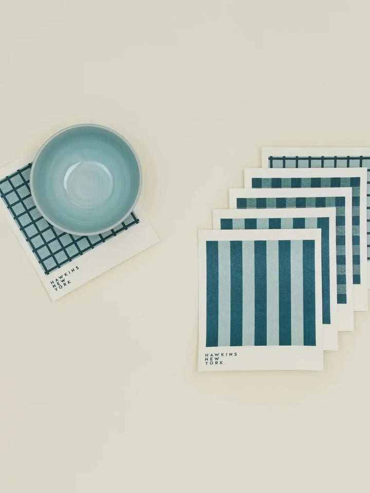 A flat lay features a light blue ceramic bowl resting on a windowpane sponge cloth alongside a stacked bundle of coordinating patterns from the Essential Swedish Dishcloth in Sky/Peacock (Set of 6) from Hawkins New York | Twentyseven Toronto
