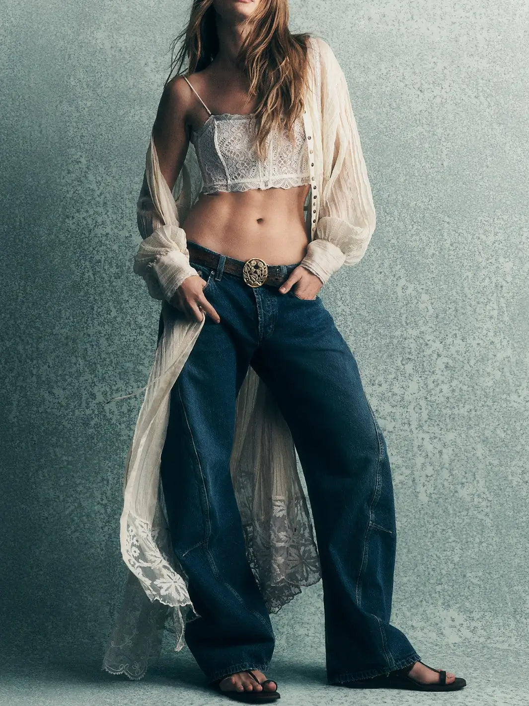 A model wearing the Free People Fable Twisted Seam Jean in Heartsong with a decorative gold buckle belt, white lace bralette, and a sheer unbuttoned shirt | Twentyseven Toronto