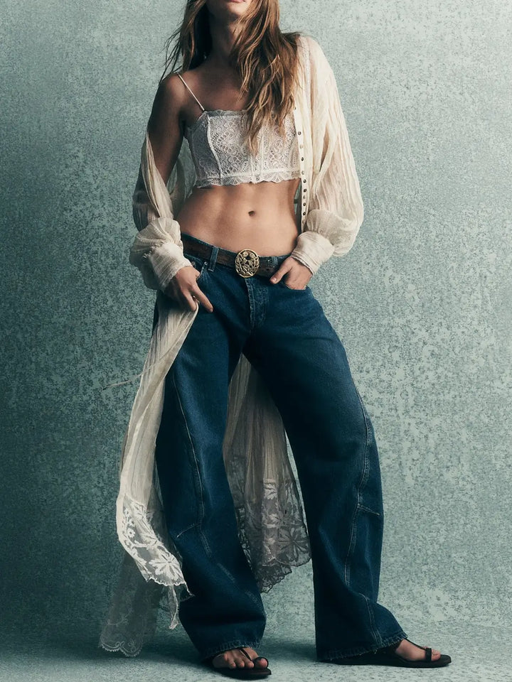 A model wearing the Free People Fable Twisted Seam Jean in Heartsong with a decorative gold buckle belt, white lace bralette, and a sheer unbuttoned shirt | Twentyseven Toronto