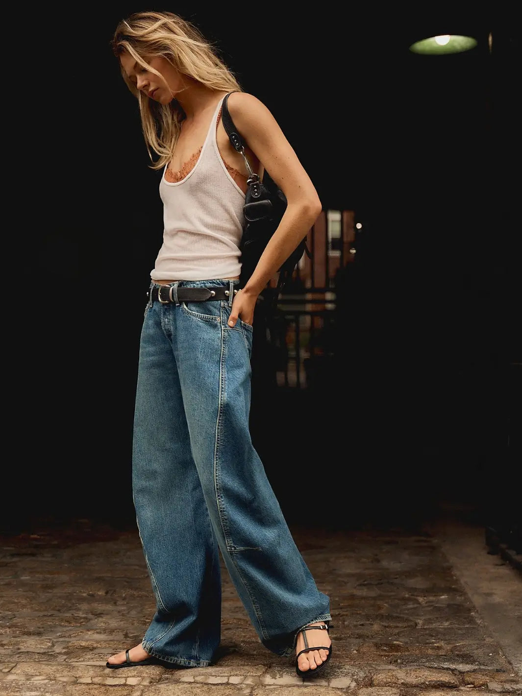 An outdoor lifestyle shot shows a model walking in the slouchy Free People Fable Twisted Seam Jean in Heartsong styled with a white ribbed tank top and black strappy sandals | Twentyseven Toronto