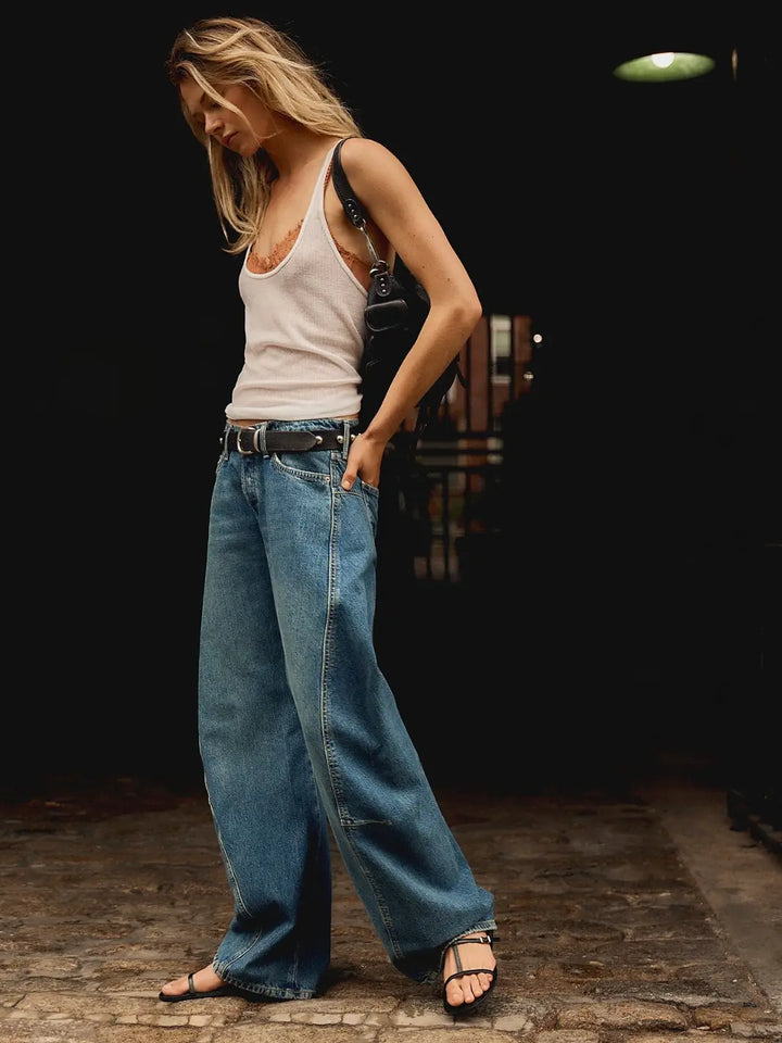 An outdoor lifestyle shot shows a model walking in the slouchy Free People Fable Twisted Seam Jean in Heartsong styled with a white ribbed tank top and black strappy sandals | Twentyseven Toronto