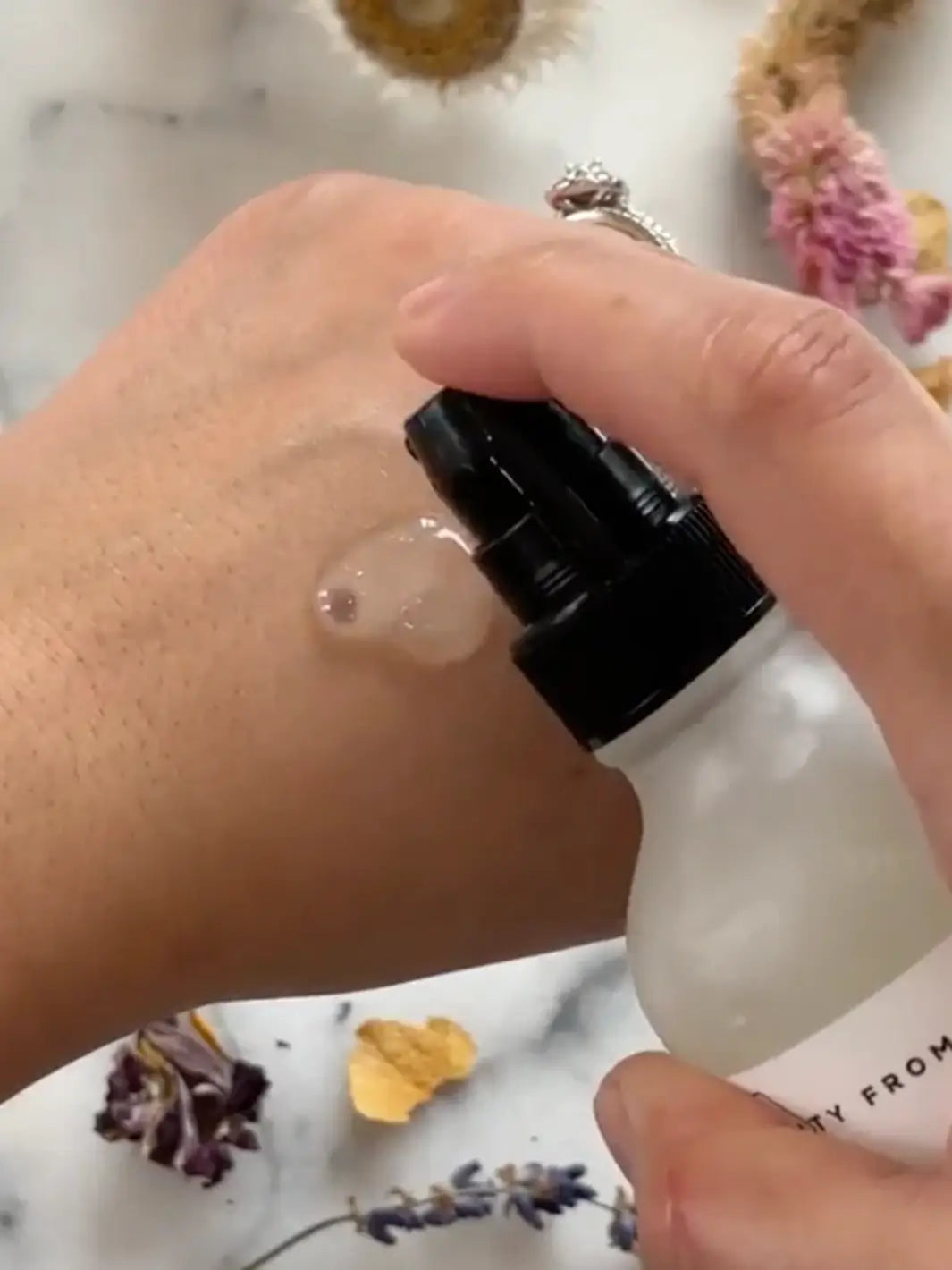 Hand holding a white bottle with black cap, applying Beauty from Bees Face Cleanser to skin against a marble background | Twentyseven Toronto