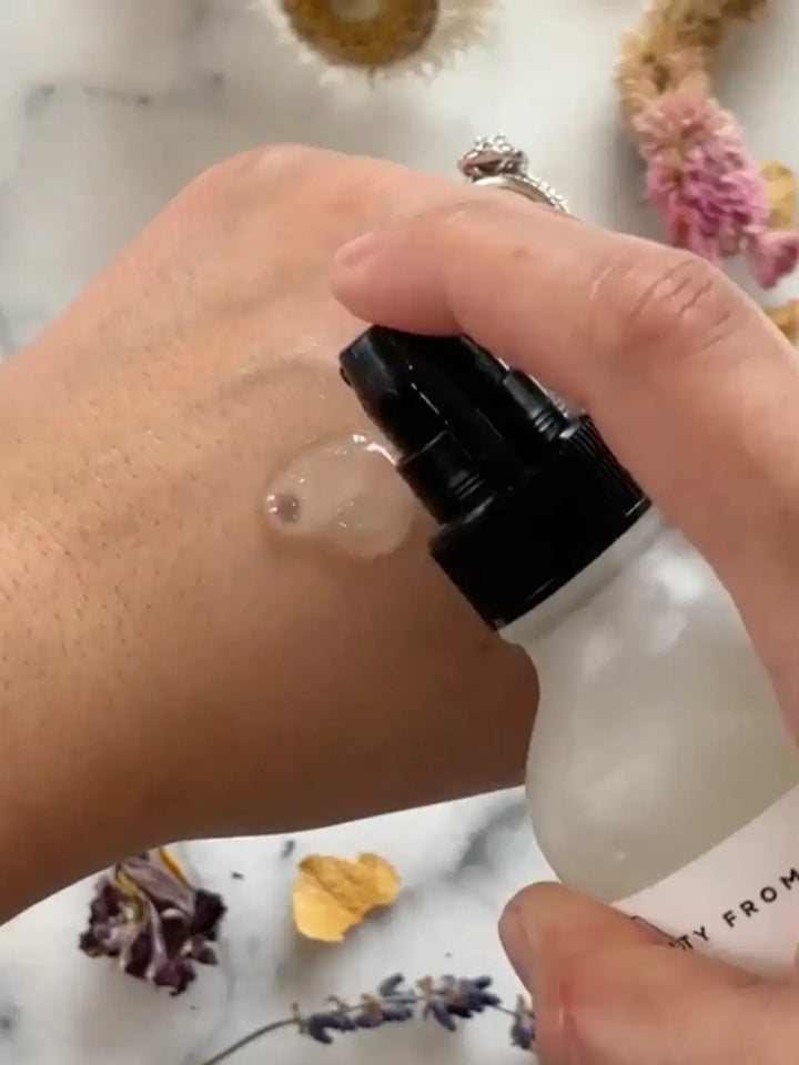 Hand holding a white bottle with black cap, applying Beauty from Bees Face Cleanser to skin against a marble background | Twentyseven Toronto