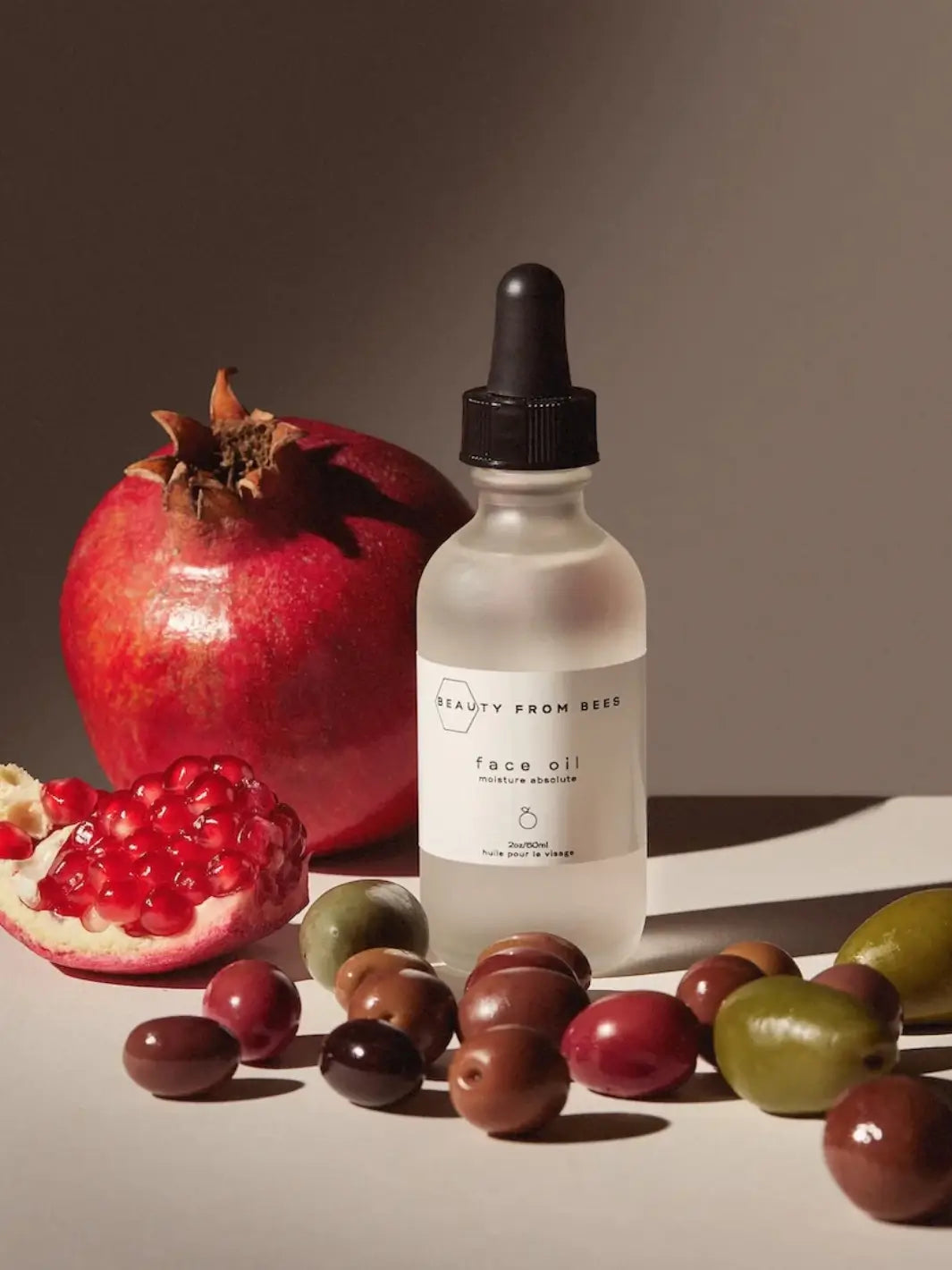 Bottle of face oil by Beauty from Bees with a black dropper surrounded by pomegranates and olives on a neutral background | Twentyseven Toronto