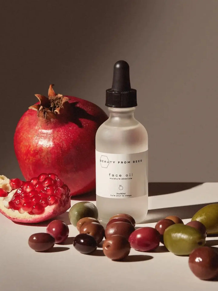 Bottle of face oil by Beauty from Bees with a black dropper surrounded by pomegranates and olives on a neutral background | Twentyseven Toronto