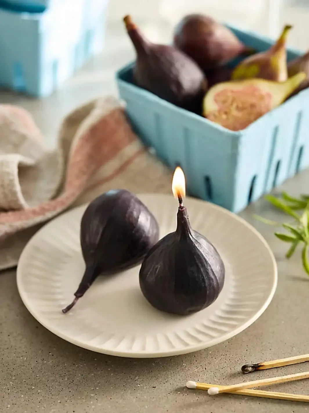 Two fig-shaped candles from Happy Organics on a white plate with various fruit shaped candles in a blue carton in the background on a table | Twentyseven Toronto