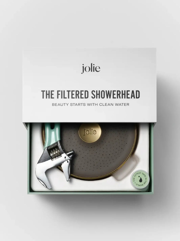 An open white and green branded box displaying the unboxed Jolie Skin Co The Filtered Shower Head - Brushed Gold alongside an included installation wrench and a small roll of plumber's tape | Twentyseven Toronto