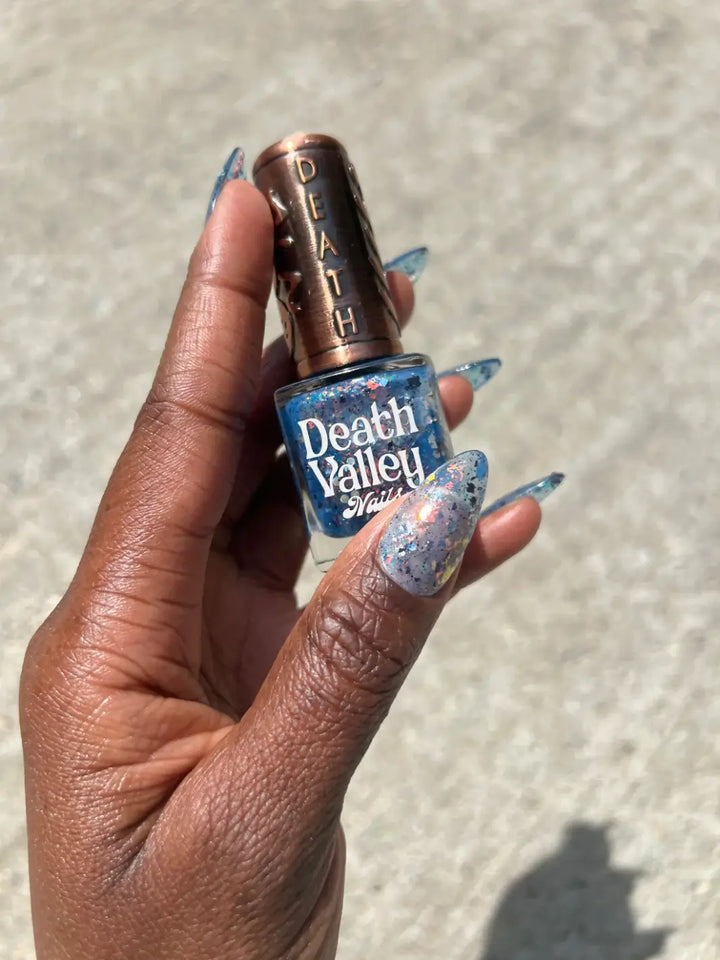 A hand with long nails showcasing the sparkling, deep blue multi-colored flakies and sheer coverage of Death Valley Nails Fish Scales Nail Polish | Twentyseven Toronto