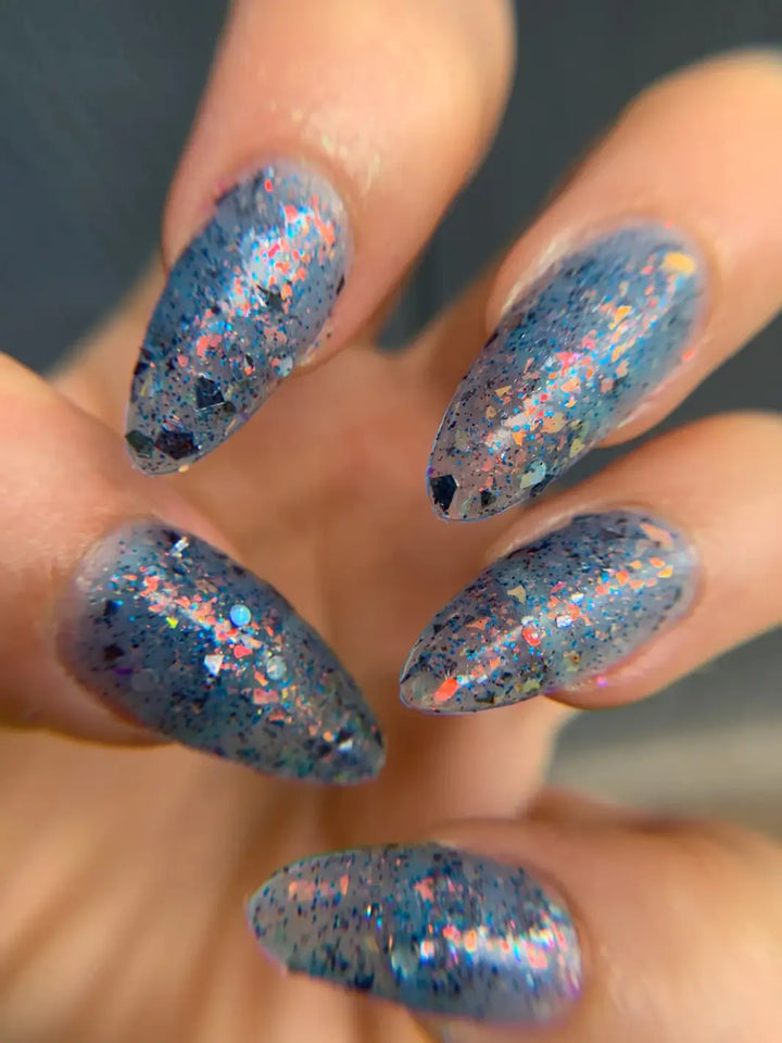 A close-up shows long, almond-shaped nails painted with Death Valley Nails Fish Scales Nail Polish, catching the light to reveal multi-colored iridescent flakes and dark specks distributed throughout the translucent blue formula | Twentyseven Toronto
