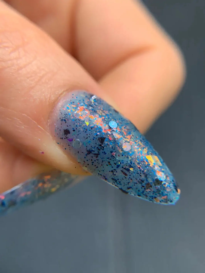 An extreme close up focuses on a single stiletto fingernail coated in Death Valley Nails Fish Scales Nail Polish, highlighting its sheer dusty blue jelly base packed with iridescent orange-to-gold flakies and dark glitter bits | Twentyseven Toronto
